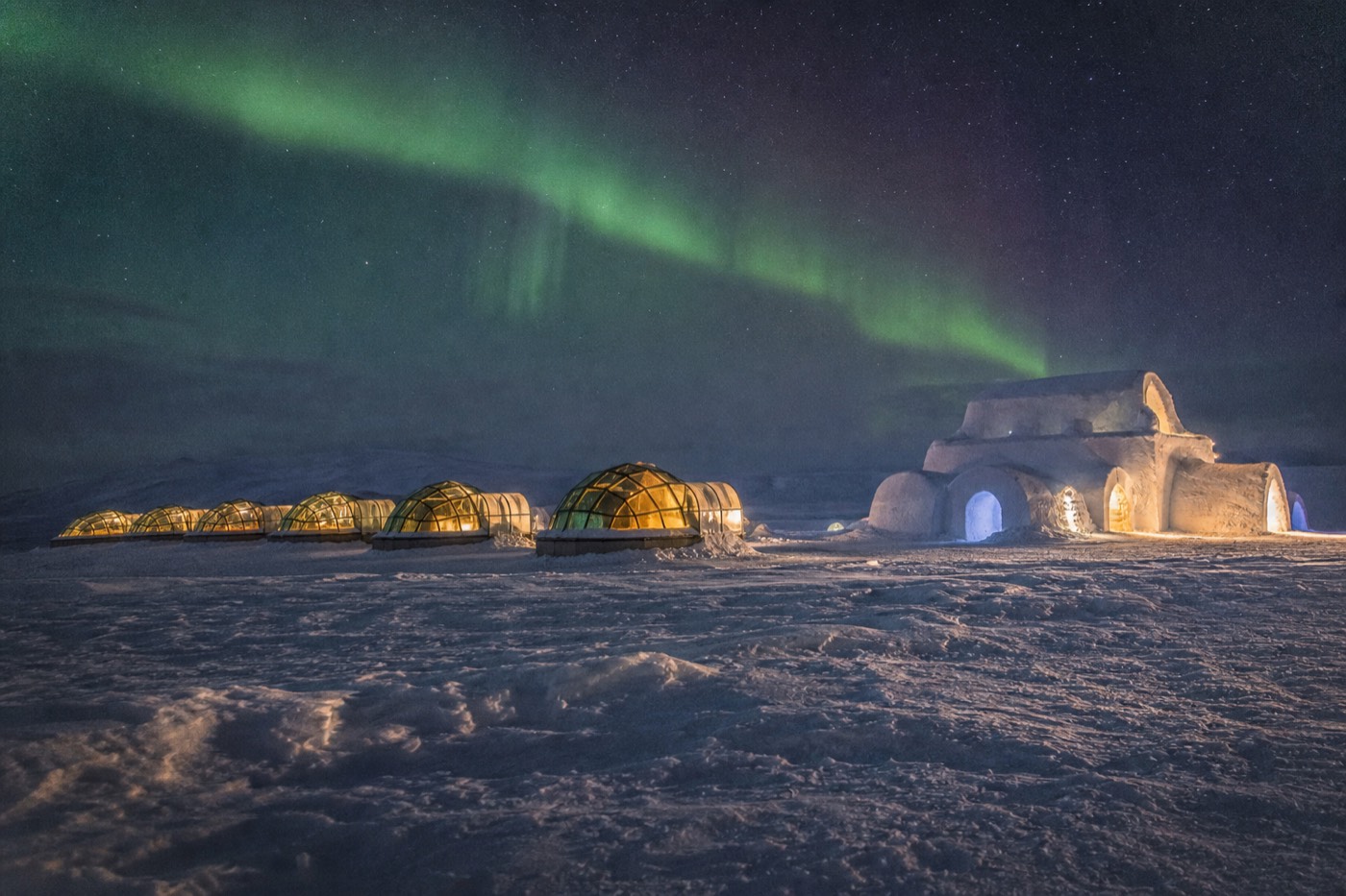 A heated glass igloo cabin in Finnish Lapland with a curved glass roof reflecting a starry sky and faint green aurora above the snow.