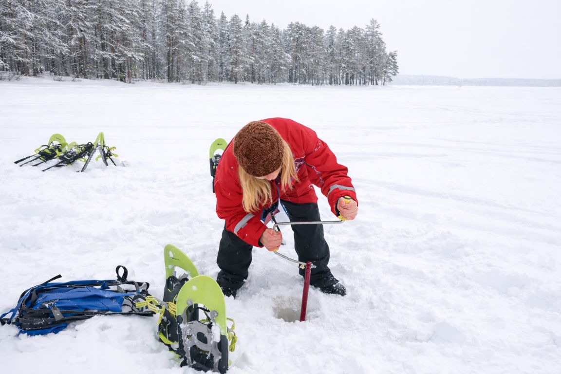 Traveler drilling hole for ice fishing on frozen lake in Lapland Finland surrounded by snowy forest, photographed during a Rovaniemi Tours excursion
