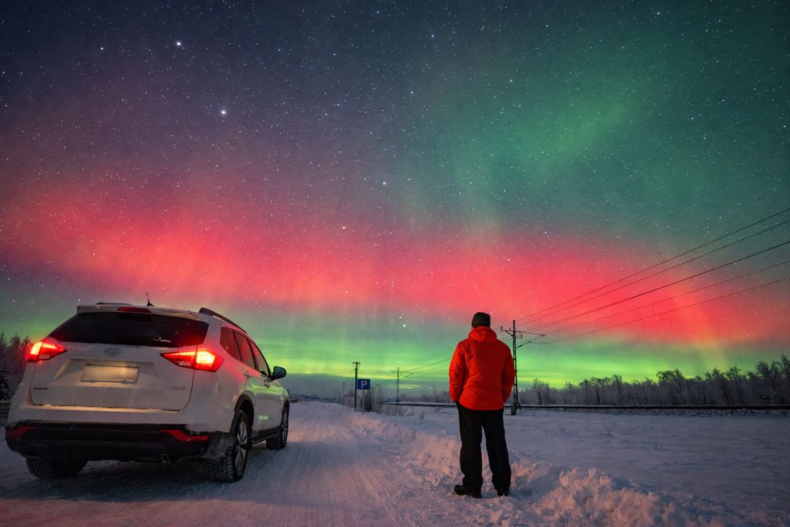 Traveler watching Northern Lights near parked car on snowy road in Rovaniemi Lapland, captured during a tour with Rovaniemi Tours
